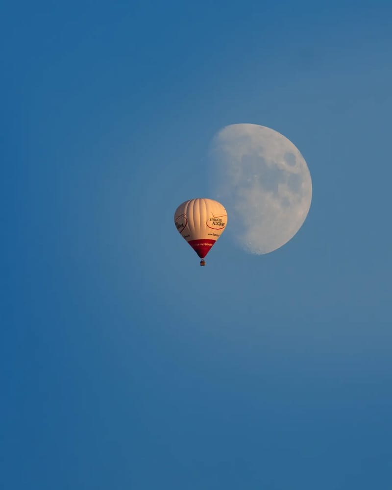 Hot air balloon with red accents floating past a gibbous moon in a clear blue sky